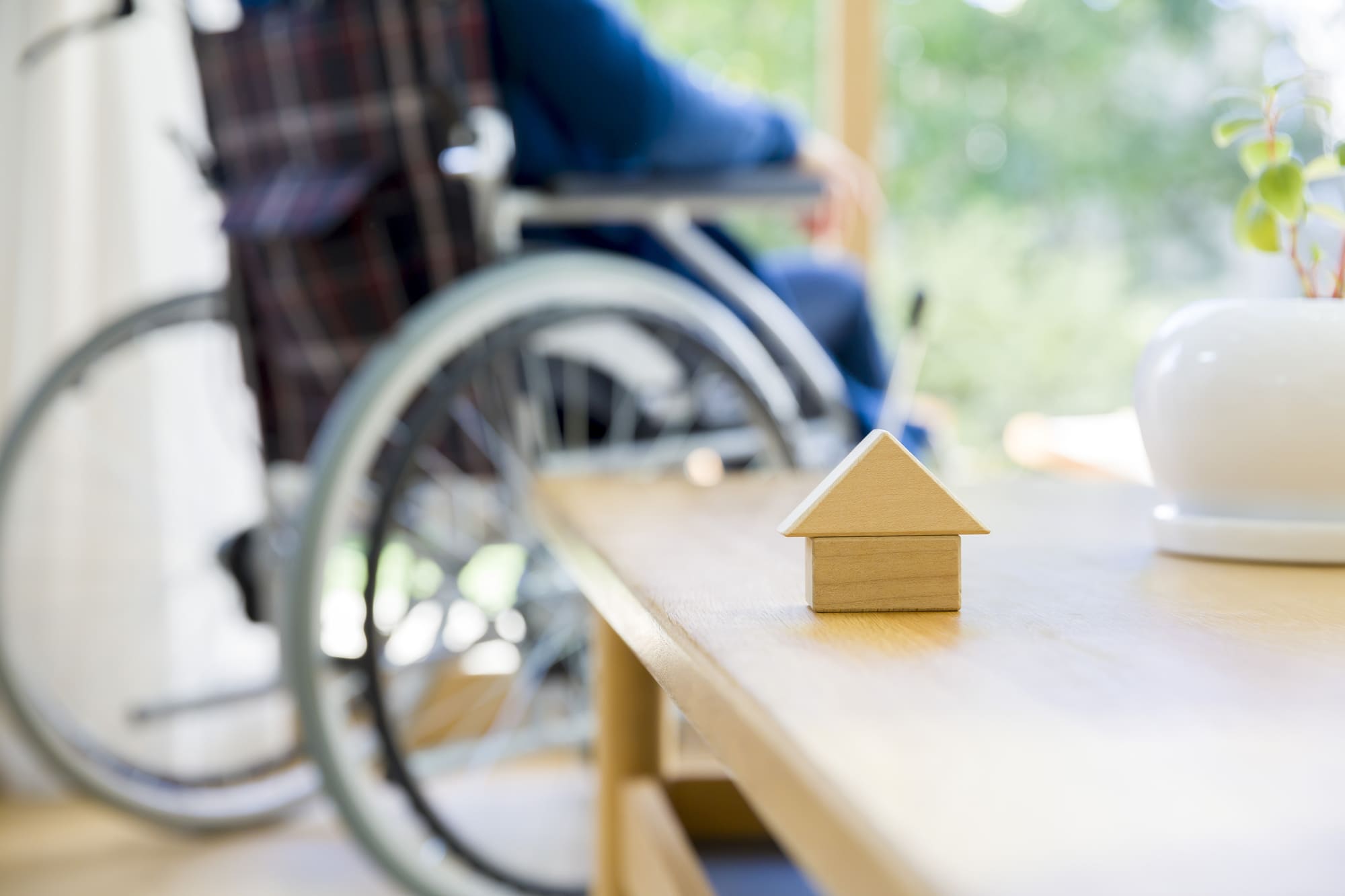 家の模型が置かれたテーブル越しに車いすの人物が見える明るいリビング